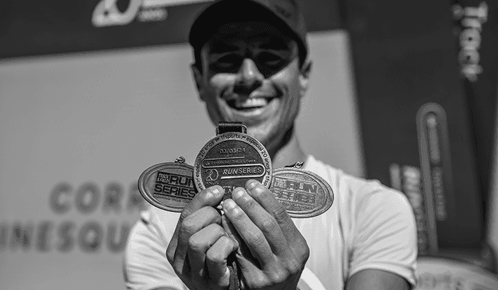 Atleta segura medalhas em primeiro plano sorrindo ao fundo. Imagem em tons de cinza. O homem está sorrindo e usando bone com aba que faz sombra em seu rosto. Nas medalhes pode-se ler Run Series.